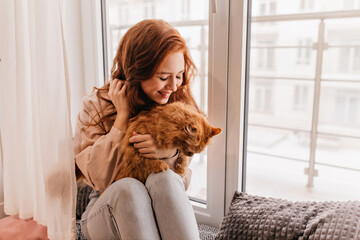 Pleasant female model holding red cat on her knees. Indoor portrait of charming ginger woman posing with pet.