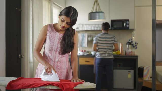 shot of a young Indian married couple doing household chores, where a wife is pressing clothes while husband is working on the kitchen