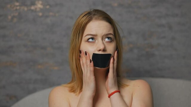 Young woman touches her taped mouth and looking at you