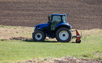 blue agricultural tractor without driver stands on a green meadow in front of a plowed field in the background, sunshine, during the day