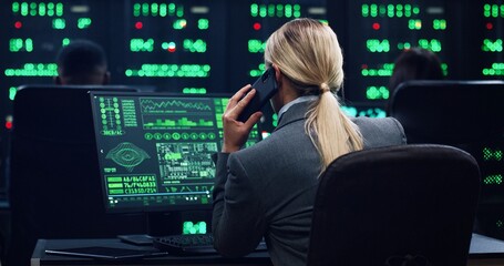 Blonde female programmer sitting at computer in server room talking on smartphone and working with database. System software concept. View from the back.