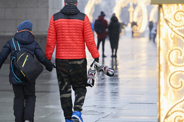  Father and son walk along the elegant alley to the ice rink. The man holds his son's hand, and in...