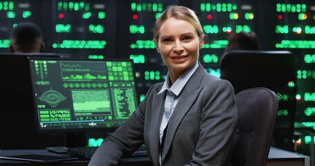 Blonde female programmer sitting at computer in server room working with database returns to camera and smiling. System software concept.