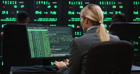 Blonde female programmer sits at computer in server room and works with database. System software concept. View from back.