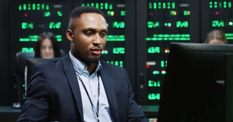 Young handsome African American programmer sitting in office at workplace on background of working servers looking at computer and taking phone call on smartphone.