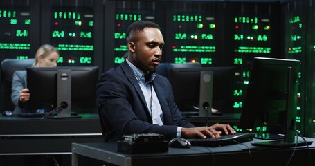 Smart African-American IT worker sitting in server room behind workplace working with database on...