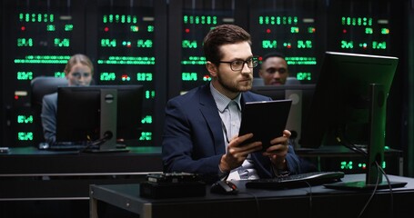 Senior IT worker with glasses sitting in server room behind the workplace holding tablet working with database on computer.