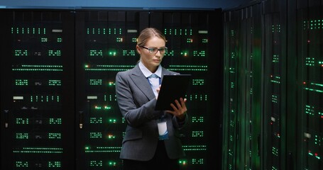 Smart beautiful female IT worker with glasses standing in server room holding tablet and working with data. Concept of system support.
