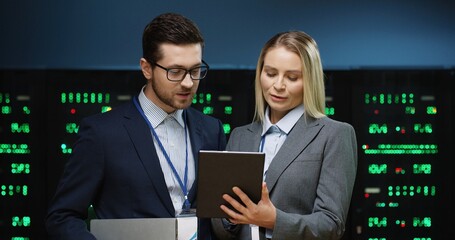 Two fellow programmers stand in server room and discuss work tasks looking at digital tablet....
