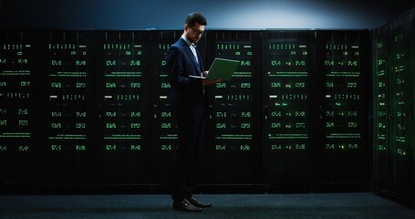Smart IT worker in glasses and suit standing in the middle of room with servers holding laptop and working with database.