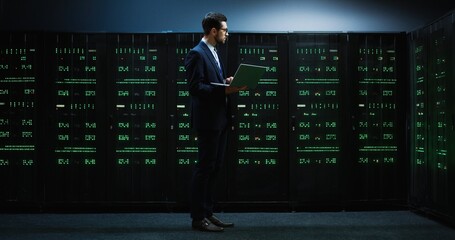 Smart IT worker in glasses and suit standing in the middle of room with servers holding laptop and working with database.
