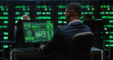 African-American IT worker sitting in server room behind workplace working with database on...