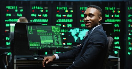 African-American IT worker sitting in server room working with database on computer returns to camera and smiling. System software concept.