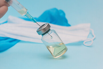 A syringe is filled with a vaccine from a jar against the background of a medical mask and blue gloves on a blue background