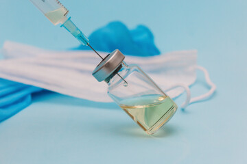 A syringe is filled with a vaccine from a jar against the background of a medical mask and blue gloves on a blue background