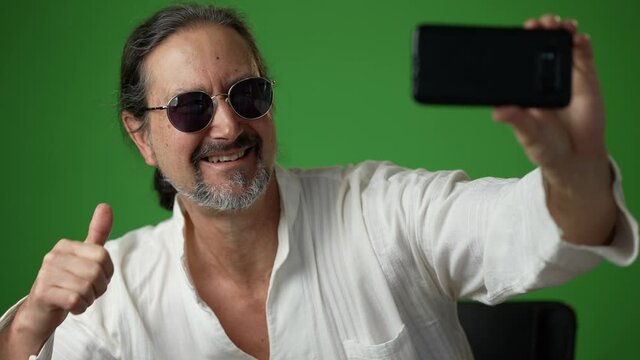 Greying man gives thumbs up, wearing sunglasses taking a selfie with a phone on a green screen chroma key background.