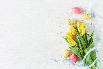 Bouquet of spring yellow tulip flowers and colorful Easter eggs top view on white wooden background.