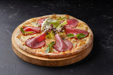 Pizza with ham several types of cheese and lettuce leaves on a dark background.
