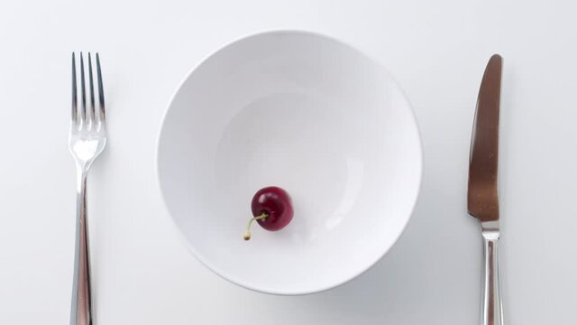 One juicy red cherry berry fall from height on a isolated empty platter with knife and fork on served laid table on white background. Table top slow motion shot, diet minimalism lunch concept.