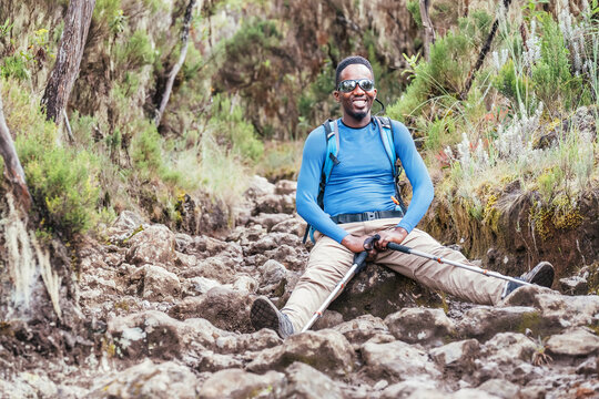 Portrait of a cheerfully smiling African-American Ethnicity young man in sunglasses sitting with backpack and trekking poles and resting on mountain trekking path. Active people and traveling concept