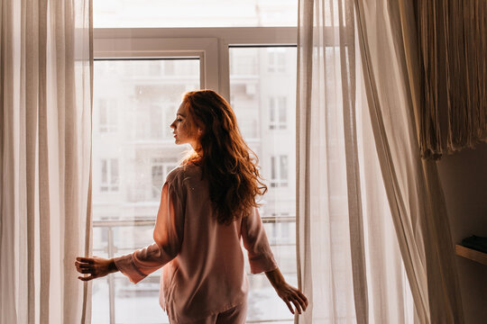Inspired long-haired woman posing in pajama. Indoor photo of serious female model standing near window in morning.