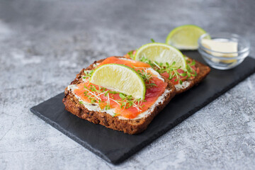 Two toast sandwiches on dark grain bread with butter, lightly salted salmon, microgreens. Idea for healthy breakfast, snack. Served on black stone board, gray background. Healthy fats omega 3