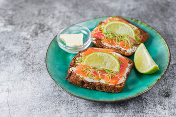 Two toast sandwiches on dark grain bread with butter, lightly salted salmon, microgreens. Served on turquoise plate, gray background with lime. Healthy fats omega 3. Idea for healthy breakfast, snack