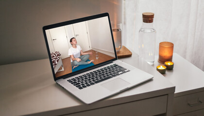 Woman meditating online at home. A video with remote yoga in a laptop's screen. A laptop with a video call on a table at home, candid atmosphere