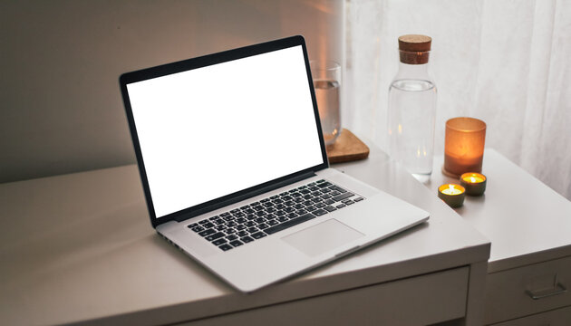 A laptop with white screen on a table with a plant, water jar, candles. Cosy atmosphere on a working place 