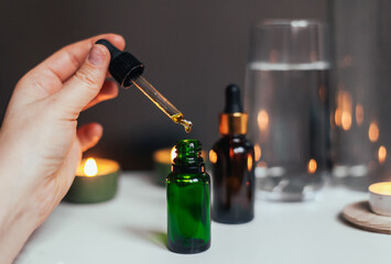 A pipette with dropping beauty oil, green and brown glass bottles, candles and water on the background