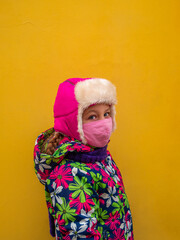 Young girl in warm winter clothes with protective pink face mask on yellow wall background. Urban...