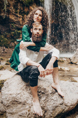 Young couple - man with beard and woman with curly hair is hugging and kissing by sitting on stone near the waterfall Slow motion