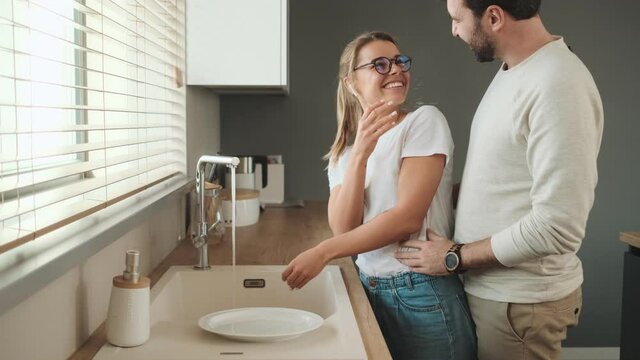 A pleased man is hugging his girlfriend while she is washing the dishes at home