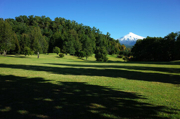 Nature of Chile. Beautiful landscape, sun and shadow, Green grass meadow and forest on hill, Snowy cone of Villarrica volcano under blue sky in sunny day. Green environment, Pucon