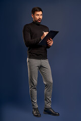 Full lenght portrait of shocked handsome bearded guy writing information to the documents on clipboard in hid hands, isolated over Pacific Blue background, copy-space.