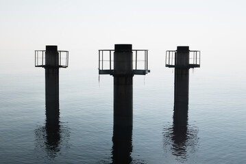 Three huge pillars of concrete emerge from the water against a strong diffused light, with...