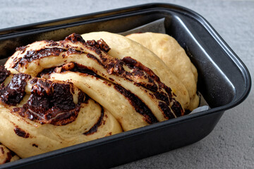 Yeast-risen braided dough cake during rising time. Unbaked krantz cake with chocolate and nuts filling in black baking tin mold.