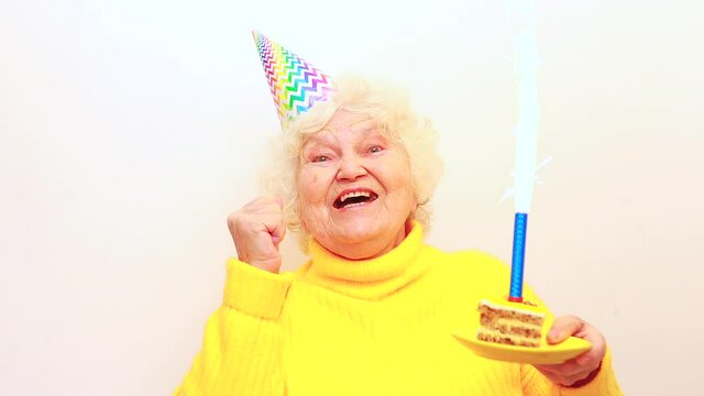 Older Woman With A Gift Wear Yellow Sweater And Horn Cap On A White Background Holding Plate With Cake With Fireworks