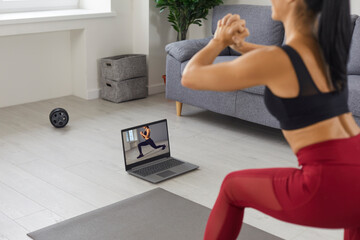 Fit woman athlete in sportswear repeating after coach making squats during online fitness workout on mat at home, rear view. Active healthy lifestyle, training at home and online training concept