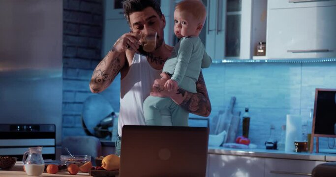Dangerous dad drinks coffee and holds the baby in his arms, preparing meals for the baby early in the morning. A tattooed man is talking on the phone drinking coffee and taking care of his son. 
