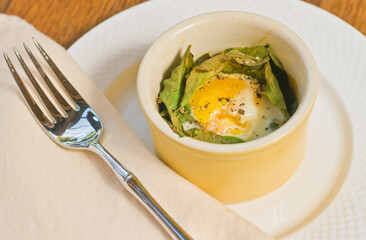 Top view, close distance of a sunny side up egg on a bed roasted kale, in yellow heat resistant bowl on  white, round plate with ar artisan fork and tan, ,linen napkin