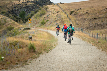 Fototapeta premium Three people cycling the Otago Central Rail Trail with Rockfall Hazard sign by the track side, at Daisybank, between Waipiata and Middlemarch, South Island
