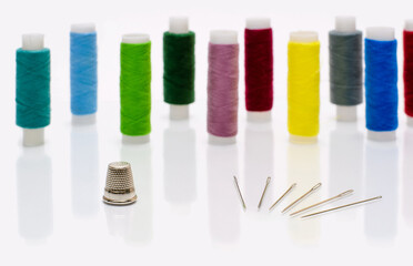 A thimble and a few sewing needles on a white glass background. In the background there are spools of thread. Selective focusing on the thimble.
