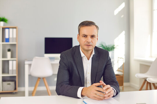 Serious Business Man Sitting At Office Desk, Looking At Camera And Listening To You. Portrait Of HR Manager, CEO, Bank Advisor, Financial Consultant, Political Expert, Teacher Or University Professor