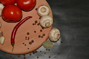 red tomatoes, mushrooms, garlic, red hot pepper, allspice on round wooden board on dark background. Top view. Space for text