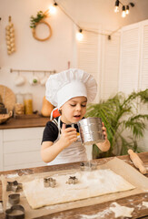 a cheerful little cook boy in a cap and apron sifts flour through a sieve in a wooden kitchen