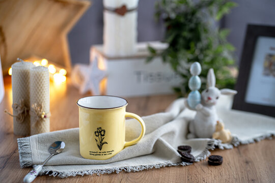 on the table there is a tea composition consisting of a yellow tin cup, a teaspoon, rows of Easter hare with Easter eggs, in the background a picture and candles. 