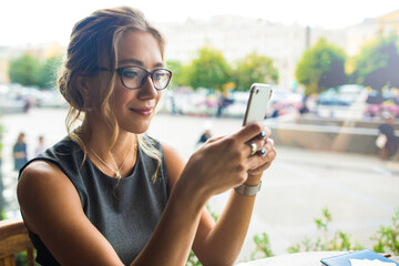 Beautiful female in glasses reading good news in e-mail on mobile phone while relaxing in restaurant