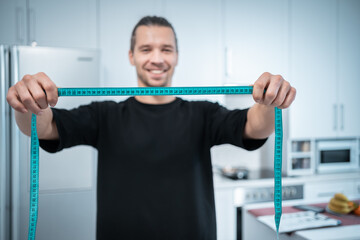 happy attractive fitness nutritionist man with measuring tape at modern kitchen