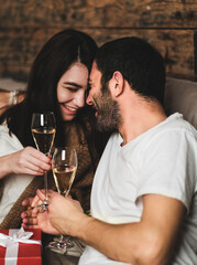 Young smiling happy couple in love in comfortable home clothing holding glasses of champagne, looking at each other and celebrating holiday at home. Valentines day celebration concept